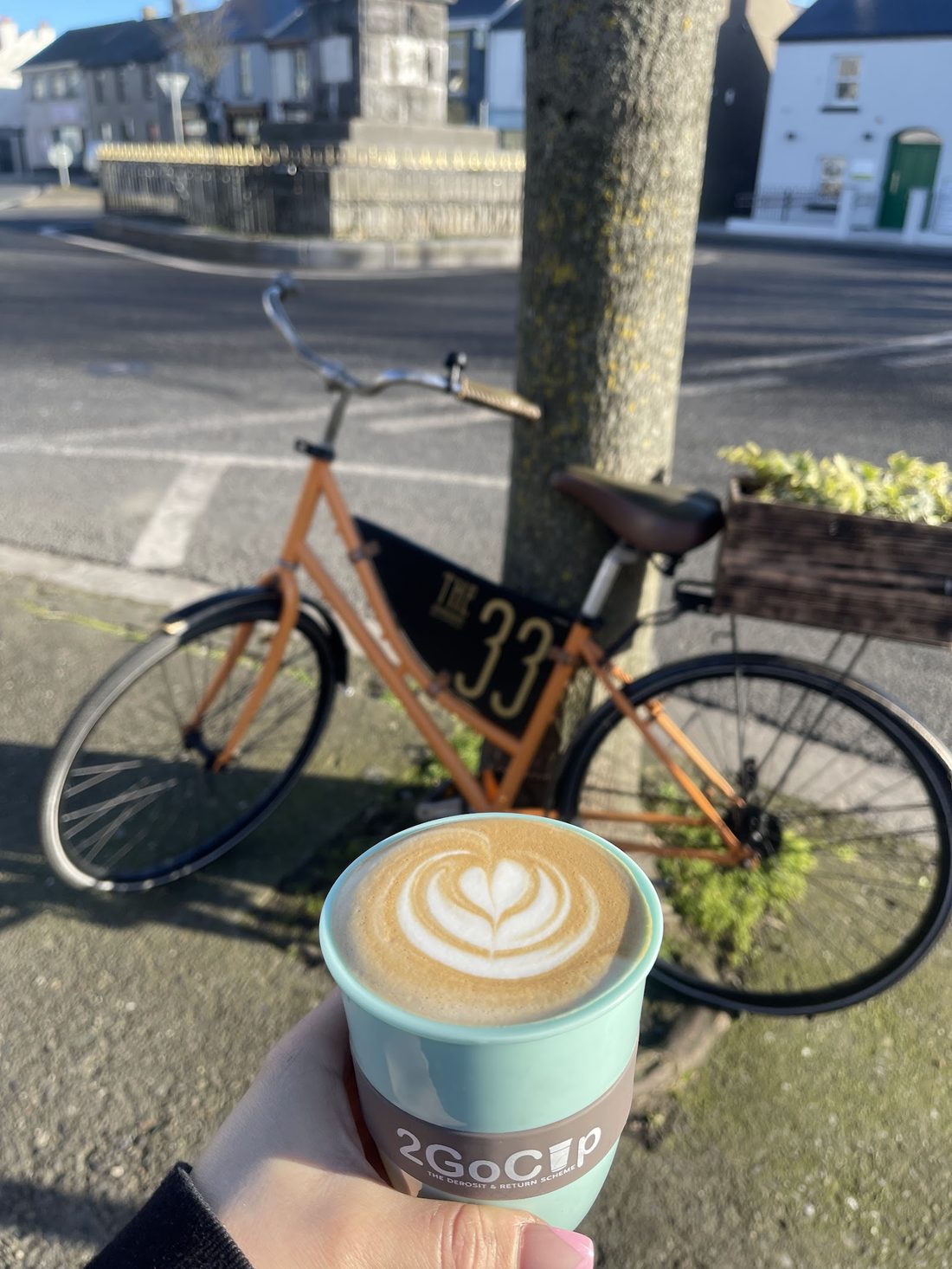 A flat white with latte art held up in front of The 33 Cafe's orange bicycle, with a "THE 33" plate, on the street in Skerries