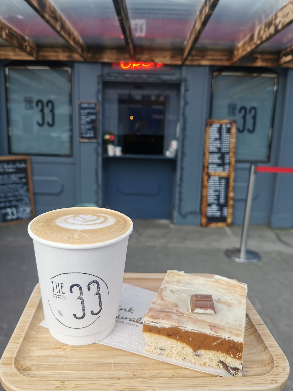 The dark timber kiosk of The 33 Cafe on Strand Street, Skerries, with chalkboard menus either side of the hatch and a branded takeaway cup and slice of cake in the foreground